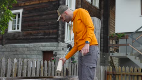 Bearded-man-in-orange-hoodie-and-cap-fills-water-bottle-at-mountain-wooden-fountain-and-smiles-looking-at-camera,-slow-motion