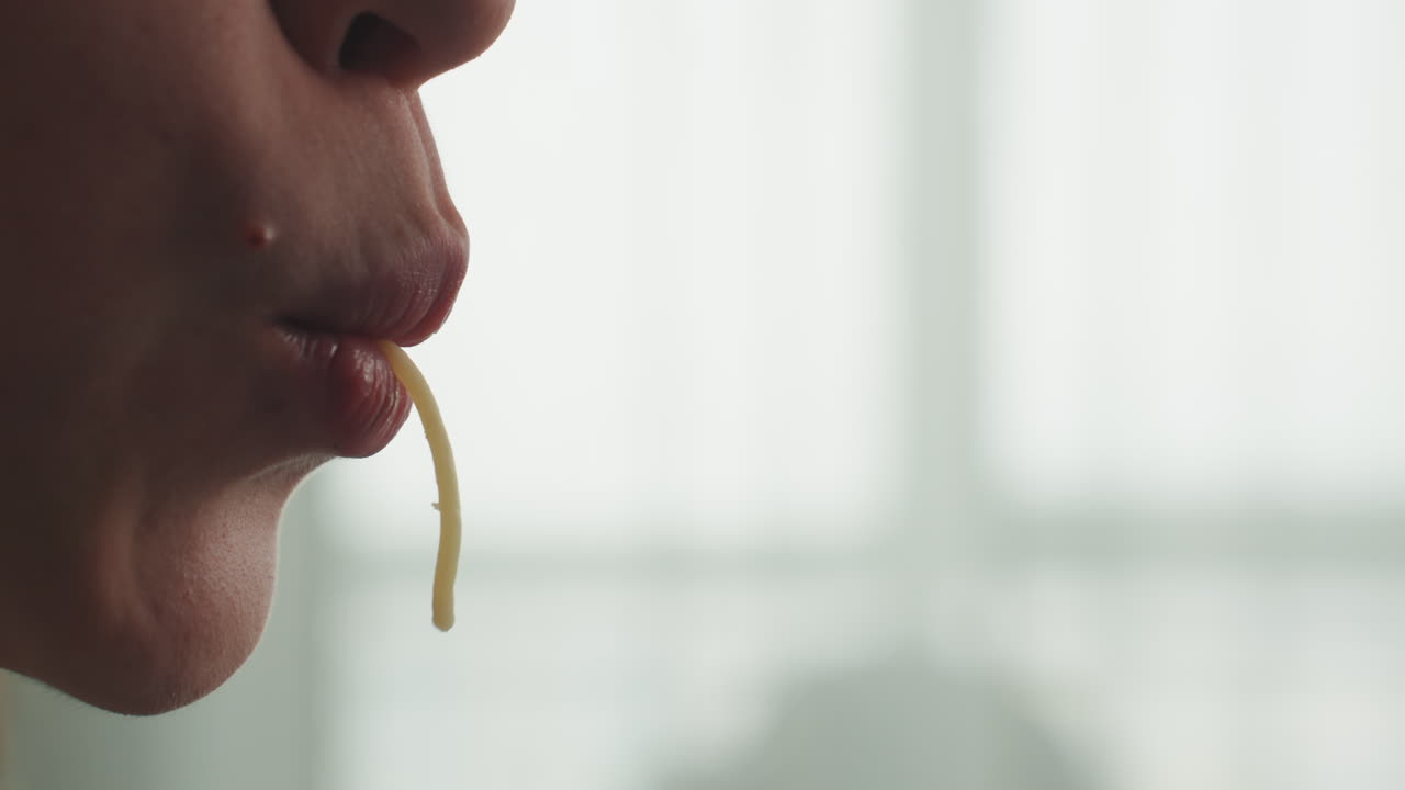 Close profile caucasian woman eating spaghetti, backlit kitchen window setting. Camera captures noodle dangling from lips, soft natural light highlighting skin texture and subtle saliva, contemplative