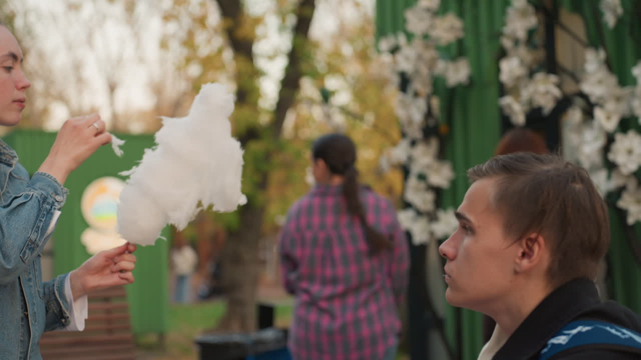 Donna caucasica al festival che offre zucchero filato a un uomo seduto tra fiori e luci, sfondo di bancarelle floreali, corteggiamento giocoso e romanticismo con sfumature matrimoniali, assaggio spontaneo, bagliore serale, affettuoso.
