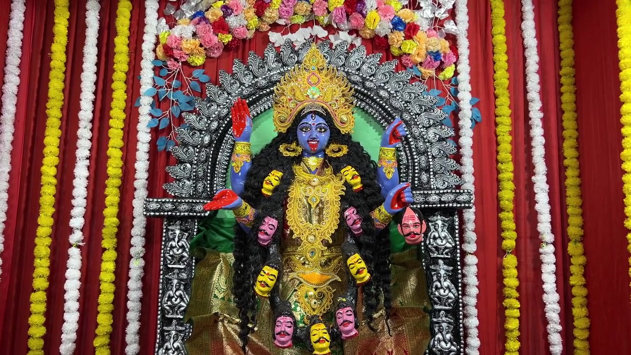 Ídolo de Maa Kali em um pandal com um fundo de tecido vermelho e guirlandas de flores amarelas e brancas.