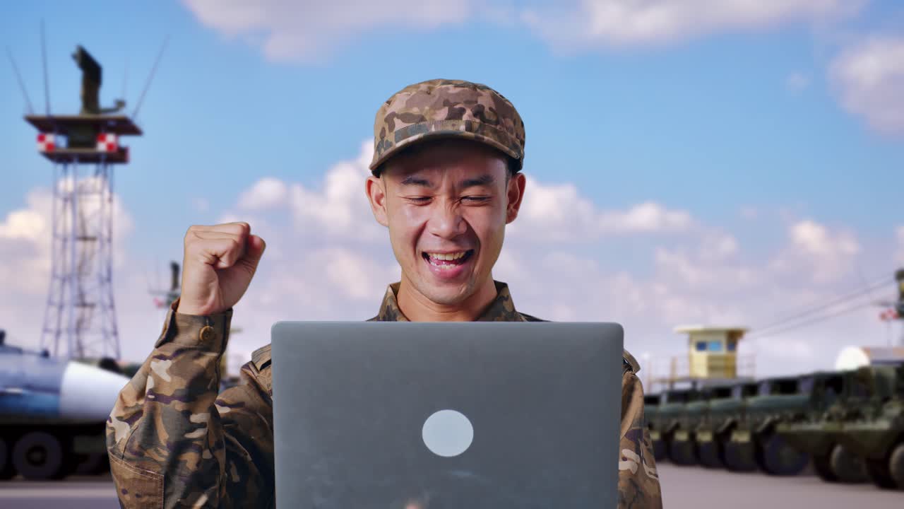 아시안 남성 군인이 노트북을 쳐다보며 소리를 지르며 군 캠프에 서서 골을 축하하고 있다.