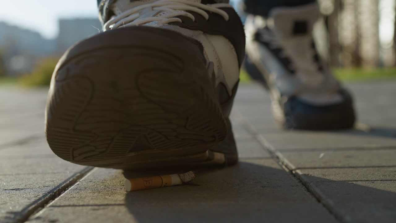 close up of burning cigarette lying on paved sidewalk with smoke swirling upward as person walks toward it and prepares to crush it underfoot