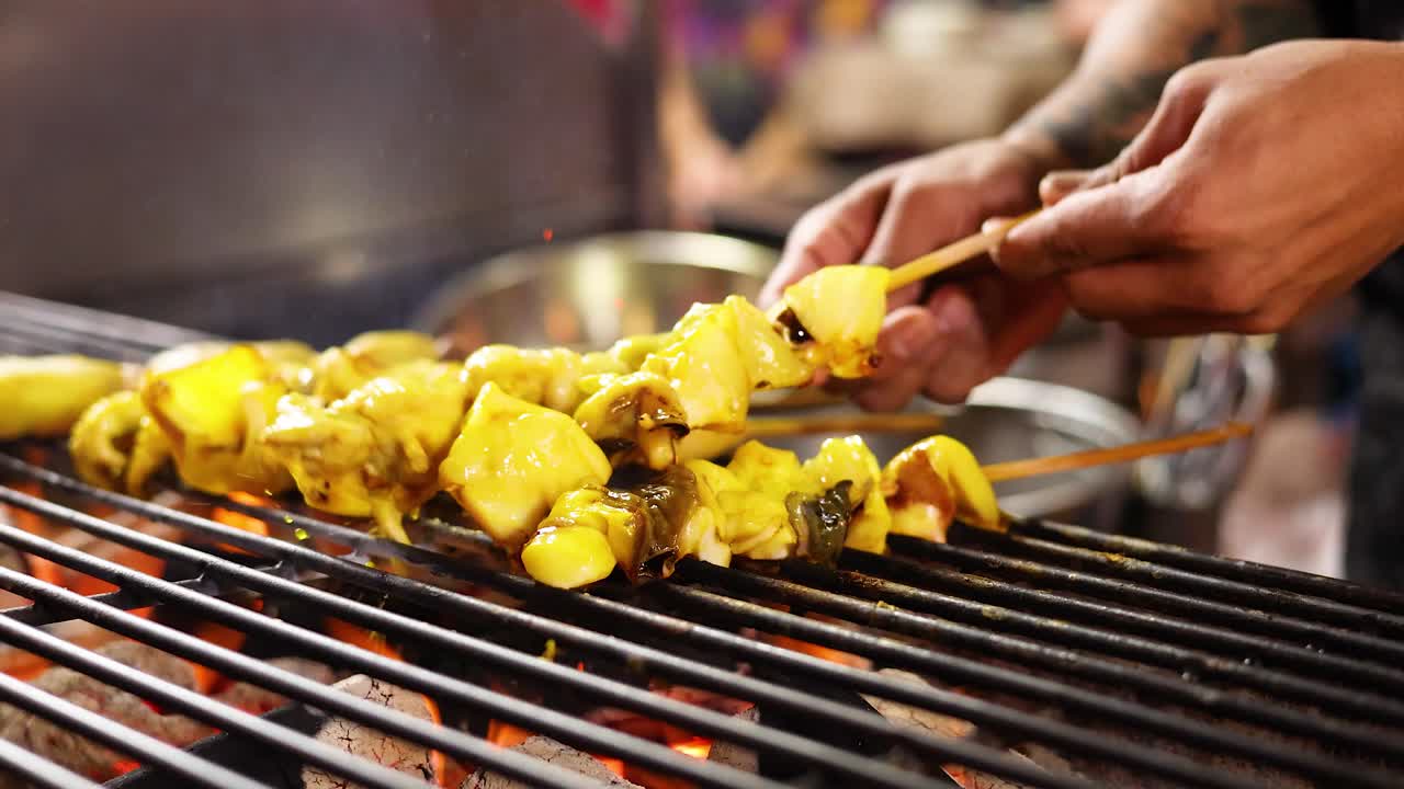 Hands grill yellow-marinated squid skewers over open flame at night market, close-up shot