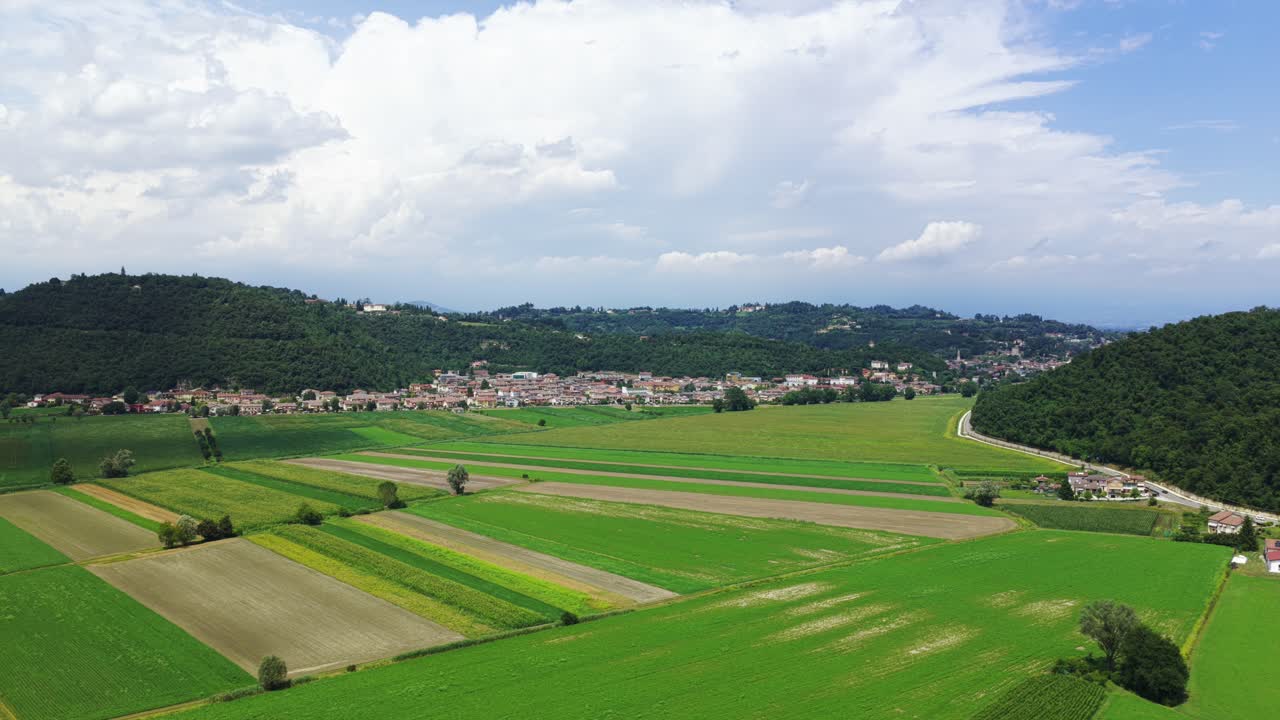 Aerial view of lush green patchwork fields in rural landscape. Village and forested hills in background under cloudy sky. For agriculture or travel content, Berici Hills, Italy