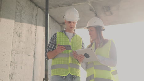 Ingenieros-Diseñadores-Un-Hombre-Y-Una-Mujer-Parados-En-El-Techo-De-Un-Edificio-En-Construcción-Y-Discuten-El-Plan-Y-El-Progreso-De-La-Construcción-Usando-Una-Tableta-Y-Un-Teléfono-Móvil.-Los-Constructores-Modernos-Discuten-La-Infraestructura-Del-Edificio-Y-Sus-Alrededores.