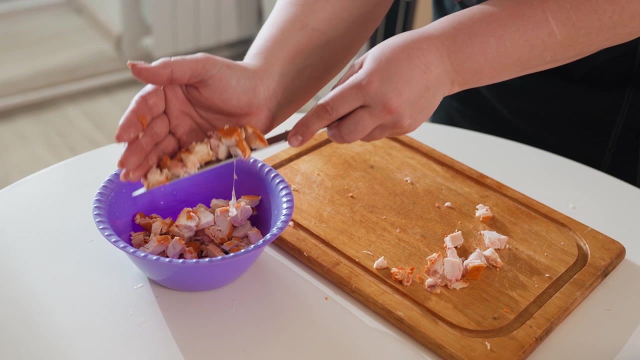 Mani adulte raccolgono prosciutto cotto a cubetti su un tagliere di legno e guidano i pezzi in una ciotola viola con un coltello e le dita, mostrando un'attenta preparazione in cucina, il trasferimento e un movimento di servizio ordinato.