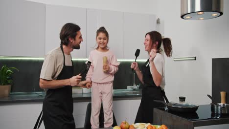 Happy-brunette-man-with-stubble-in-a-black-apron-together-with-his-brunette-wife-in-a-white-T-shirt-and-little-daughter-in-pink-clothes-dance-and-jump-while-in-the-kitchen-while-preparing-breakfast-on-a-happy-morning-in-a-modern-kitchen