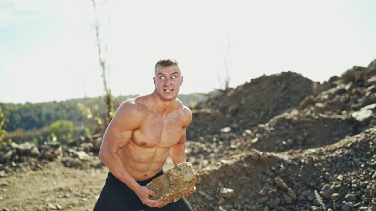 Strong athlete training his body by throwing stones. Muscular shirtless guy doing his hard workout with rocks among nature in summer. Slow motion.