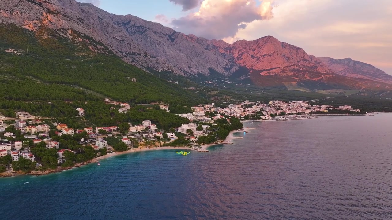 Brela, Kroatiens kustlinje visar vatten, strand och byggnader. Berg reser sig i bakgrunden. Staden sträcker sig längs kusten. Solnedgången skapar skuggor och färger i landskapet.