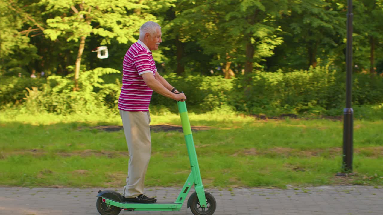 Atractivo, activo y elegante hombre mayor (abuelo) montando un patinete eléctrico en un parque, mostrando el pulgar hacia arriba.