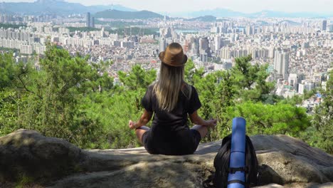 Niña-Sentada-En-La-Roca-Y-Practicando-Una-Pose-De-Loto-De-Yoga-Contra-El-Horizonte-Del-Distrito-De-Seocho-gu-Desde-La-Montaña-Gwanaksan-En-Seúl,-Corea-Del-Sur