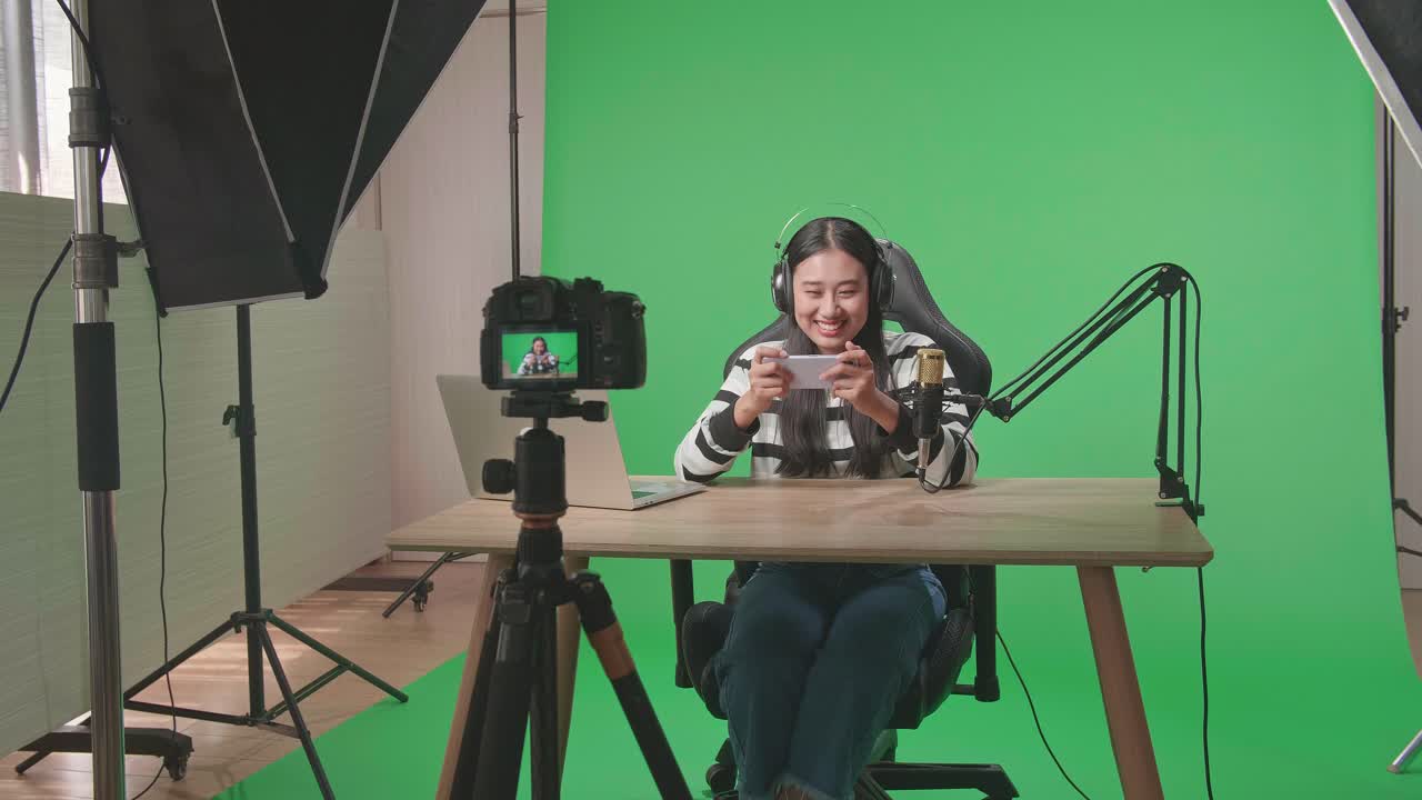 Una telecamera riprende una gamer asiatica sorridente con cuffie e computer, che guarda la telecamera mentre gioca a un gioco per cellulare su schermo verde con attrezzatura di illuminazione professionale.