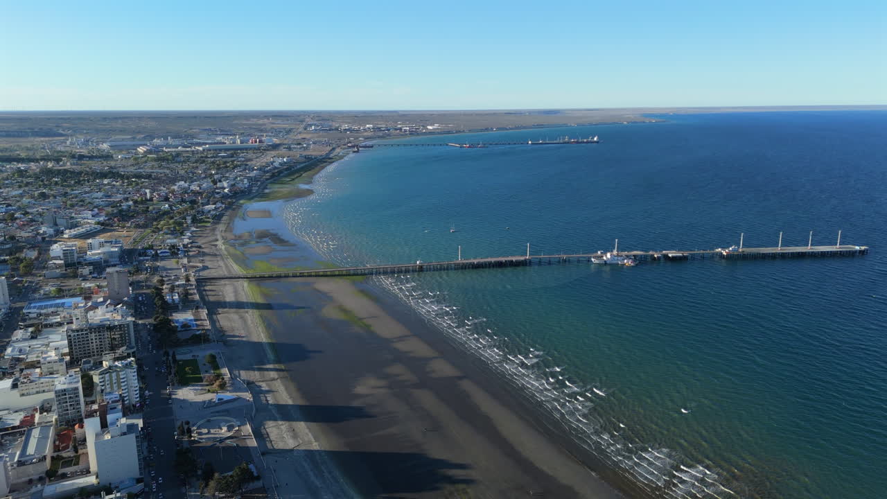 Aerial tracking left of Puerto Madryn cruising ship pier, rugged coastline and vibrant blue waters in Patagonia, establishing city