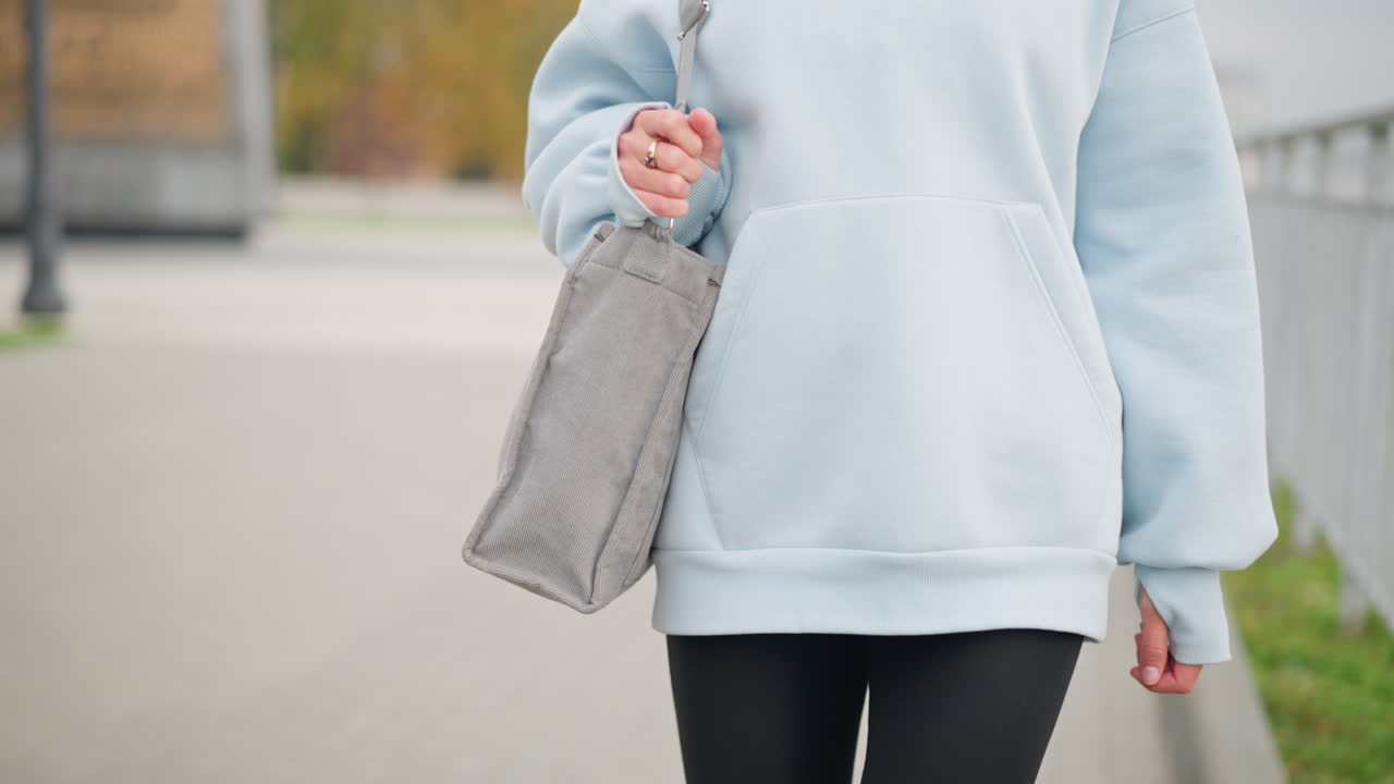 Close-up view of woman in casual clothing carrying bag with golden ring on her finger walking gently with soft steps outdoors along an iron fence and green grass path