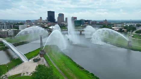 Aerial-view-of-Dayton,-Ohio:-River-with-dramatic-water-fountains,-pedestrian-bridge,-and-city-skyline-against-a-cloudy-sky-backdrop