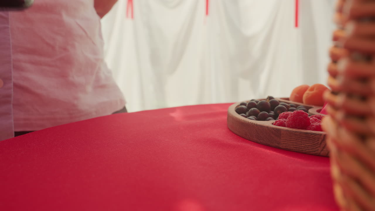 red table picnic scene with wine, bottle beside fruit platter and wicker basket, partial view of person in light shirt, white drape background, cozy intimate mood, soft ambient light, close framing