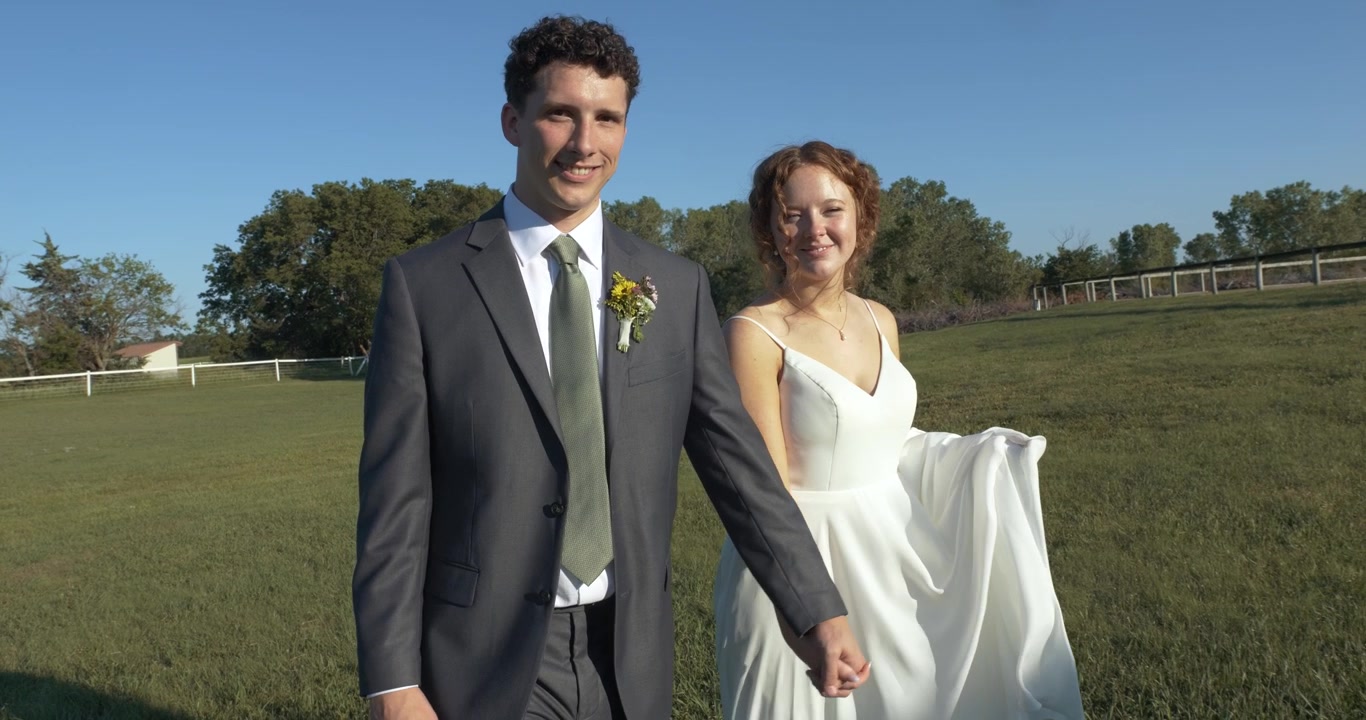 Um casal de noivos alegre caminha de mãos dadas por um campo ensolarado, compartilhando sorrisos e risadas, enquanto celebram seu dia especial em um cenário pitoresco ao ar livre cercado pela natureza
