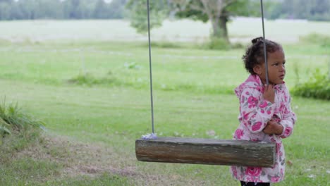 cute little african girl in pink raincoat wants to play on swing in park, static