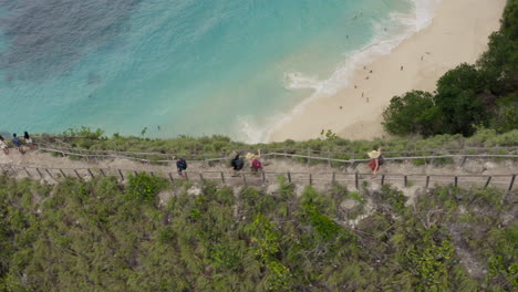 Touristen,-Die-Den-Grat-Entlang-Gehen,-Mit-Einem-Weg-Hinunter-Zum-Strand-Von-Kelingking-Unten,-Mit-Wellen,-Die-An-Land-Brechen,-Nusa-Penida,-Bali,-Indonesien