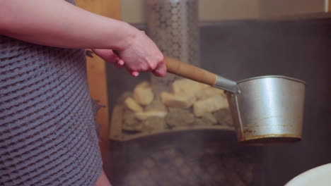close up of person pouring water from ladle onto hot sauna stones, steam rising around wooden panels, towel wrapped around waist, creating warm, calming wellness ritual in intimate spa session
