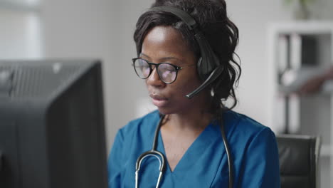 médica general negra afroamericana con bata blanca sentada en el escritorio de la oficina del médico y desplazando el ratón de la computadora mientras lee el historial médico del paciente