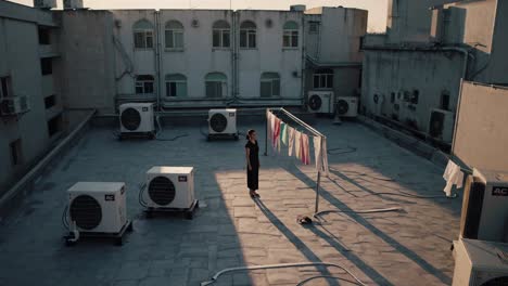 woman standing on urban rooftop during sunset, laundry hanging on clothesline, air conditioning units surrounding her, capturing peaceful city atmosphere