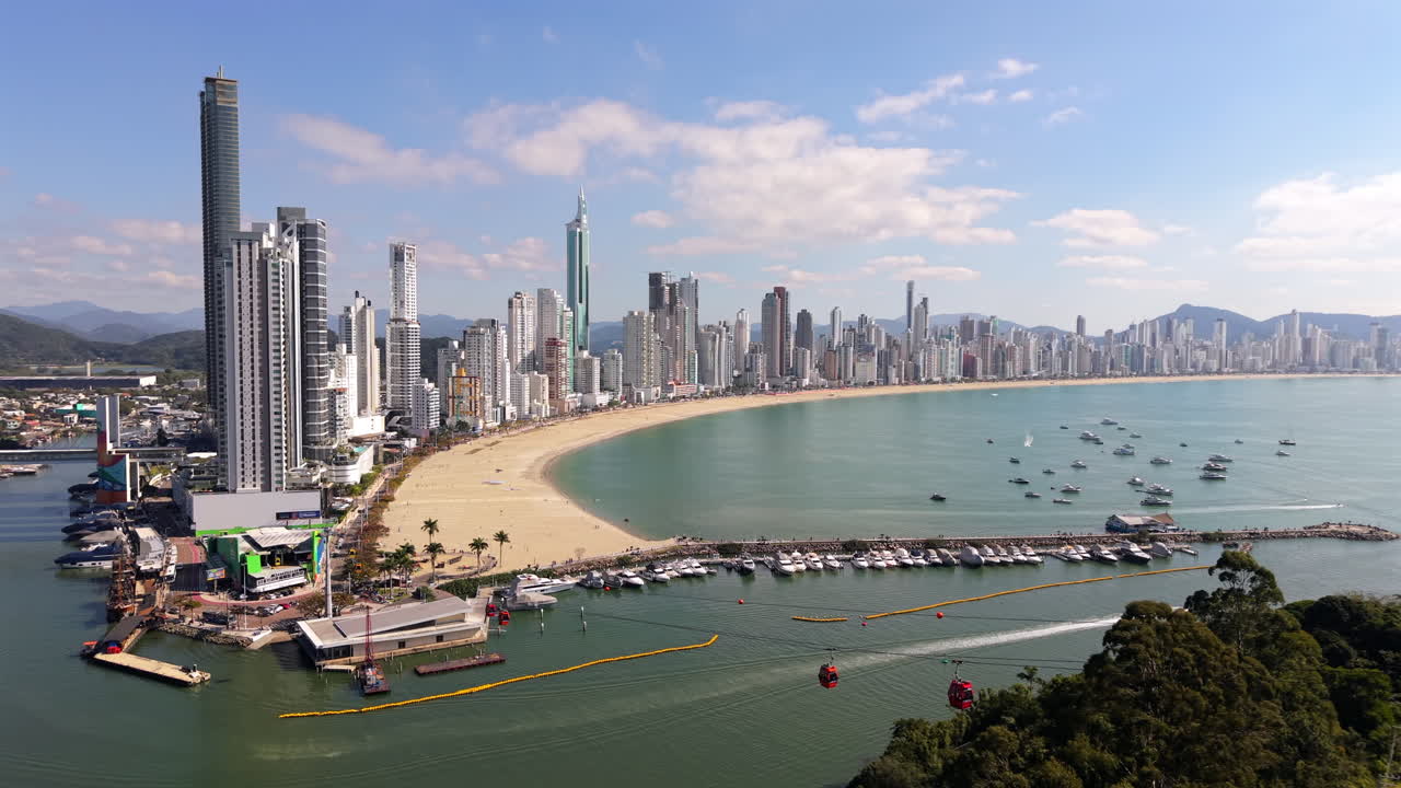 Curved beach coastline at Balneário Camboriú beside modern commercial offices and calm ocean with moving cable cars, Santa Catarina, Brazil