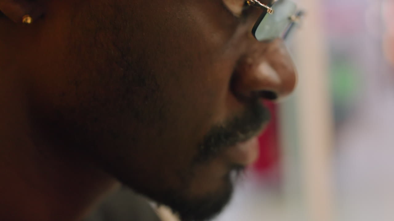 Profile closeup with gold glasses, man in soft light, earring glint, focused downward gaze, blurred colorful background, calm concentration, contemplative mood, urban cafe setting