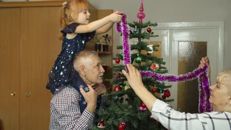 Girl-kid-with-senior-grandma,-grandpa-decorating-artificial-Christmas-tree-with-ornaments-and-toys