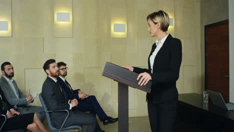 caucasian businesswoman speaking on the podium in a conference room and demonstrating video presentation on the screen behind her