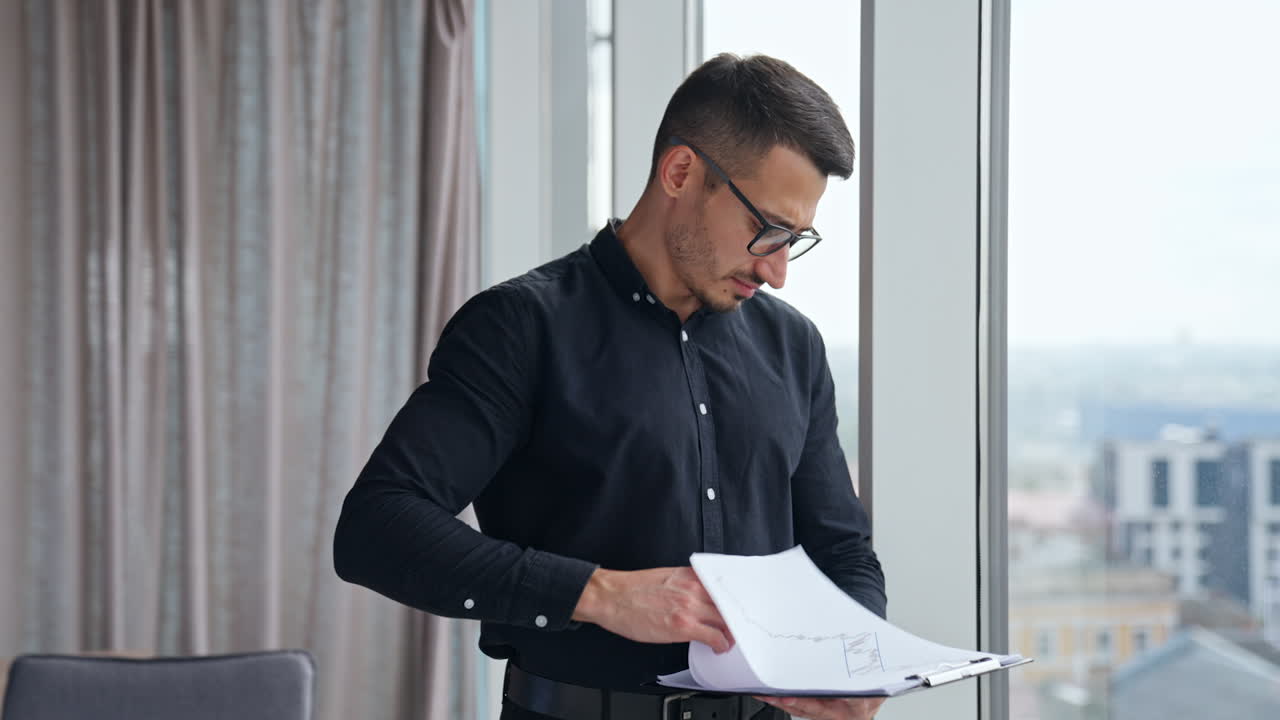 Worried man turning pages of report. Man in black clothes looks through the papers with charts with concerned expression on his face.
