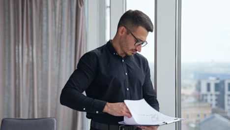 worried man turning pages of report. man in black clothes looks through the papers with charts with concerned expression on his face.