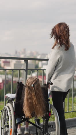 madre e hija con discapacidad miran el panorama de la ciudad desde un punto de observación en el parque. niña pequeña sentada en silla de ruedas disfrutando de un paseo con mamá