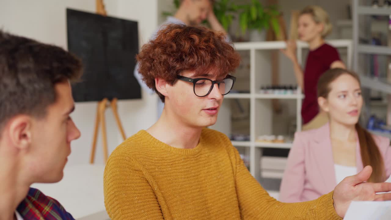 Tilt up medium shot of creative redhead young man in eyeglasses explaining his ideas to colleague sitting at table in office
