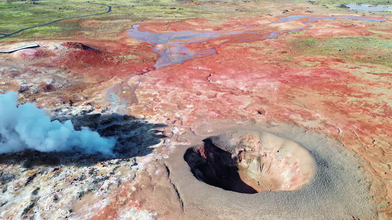 Pemandangan udara drone dari lubang panas bumi yang mengepulkan uap dan kolam lumpur mendidih di lapangan panas bumi vulkanik di Islandia. Tanah kaya mineral dalam nuansa krem, merah, dan biru mengelilingi fumarol aktif yang mengeluarkan uap.