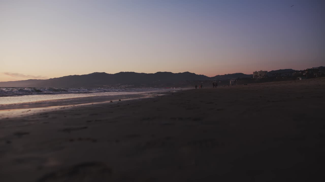 olas en la playa con gente caminando a lo lejos por la noche con montañas al fondo