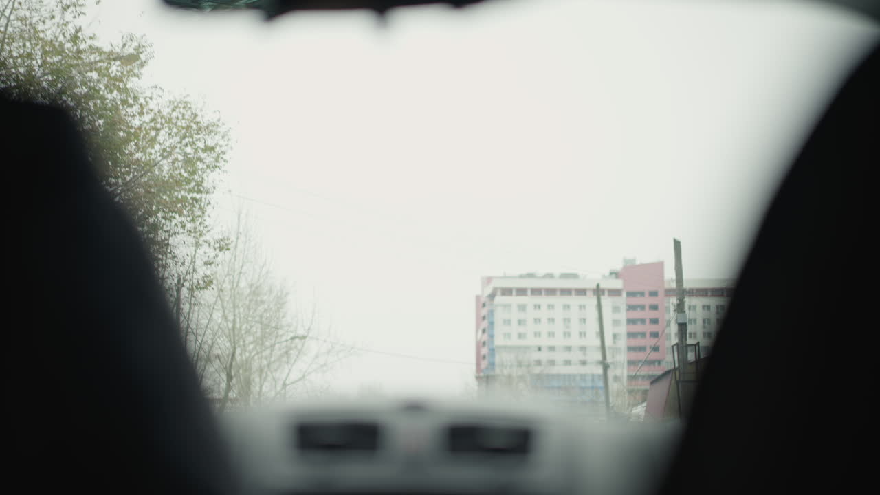 Front perspective inside moving vehicle passing leafless winter trees tall city buildings under overcast sky with blurred motion through car interior window revealing silhouette of seats