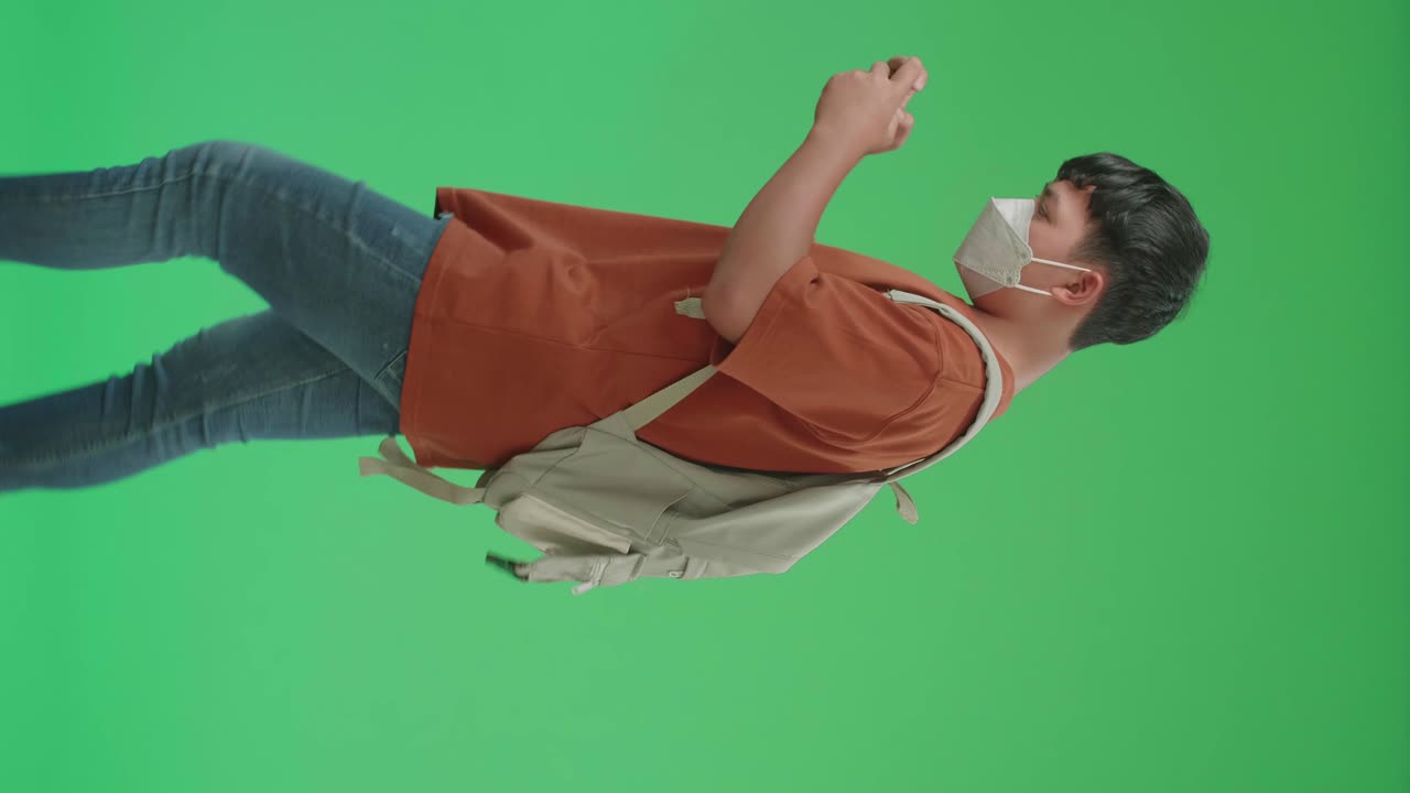 The Side View Of Asian Boy Student Wearing A Mask, Backpack, T-Shirt, And Jeans Playing Game On Mobile Phone While Walking To School On Green Screen Chroma Key.
