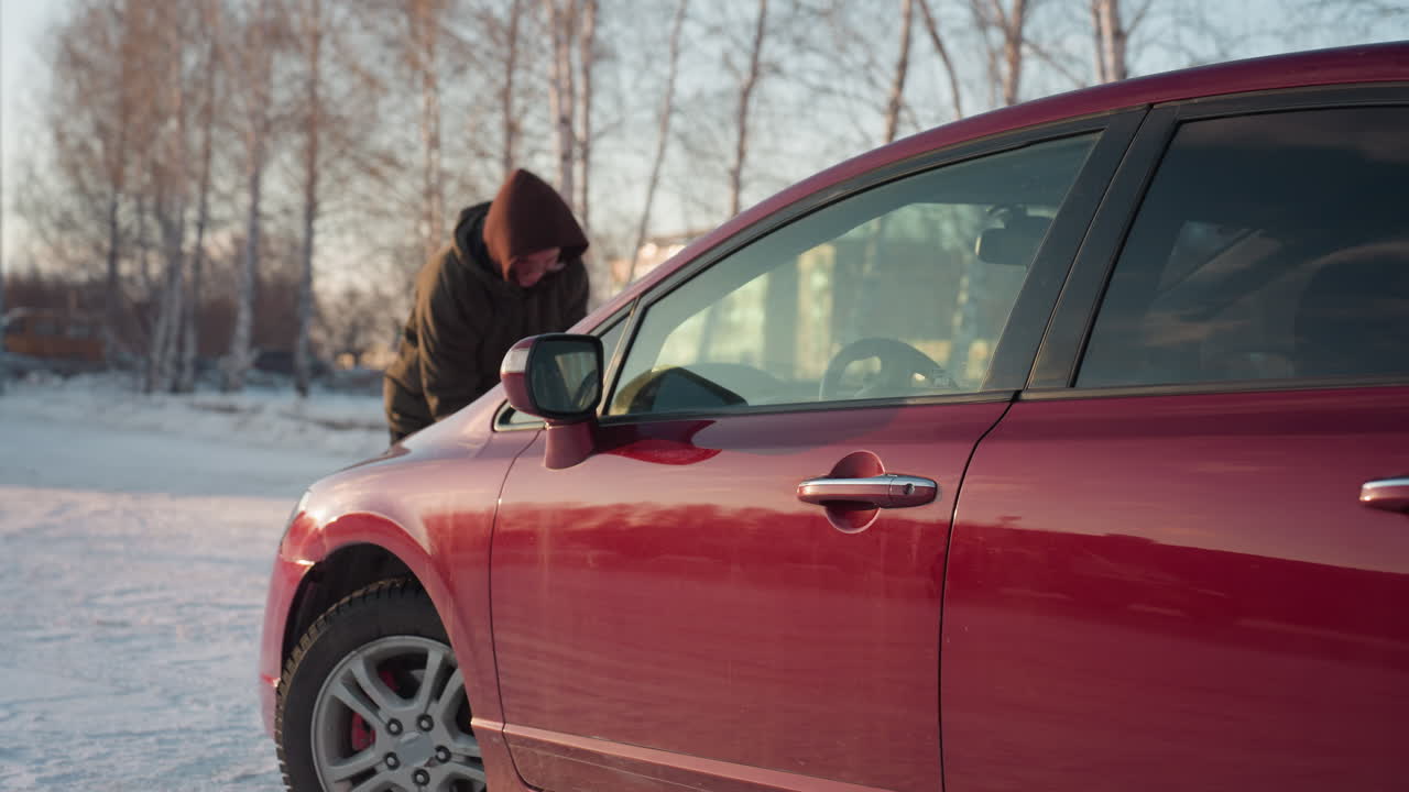 Young man wearing hoodie leans over front of parked red car in snowy outdoor lot, attempting to open bonnet with winter trees and building in background during sunny cold day