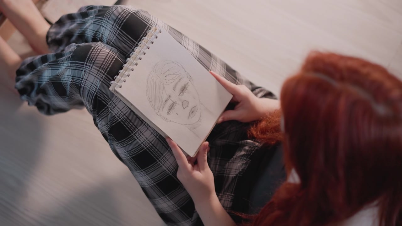 Donna caucasica dai capelli rossi che studia una scheda viso, con un ritratto su un quaderno da disegno in grembo, tracciando con cura i lineamenti con il dito. Dettagli a grafite, pantaloni a quadri su un pavimento di legno, in una pratica creativa focalizzata.