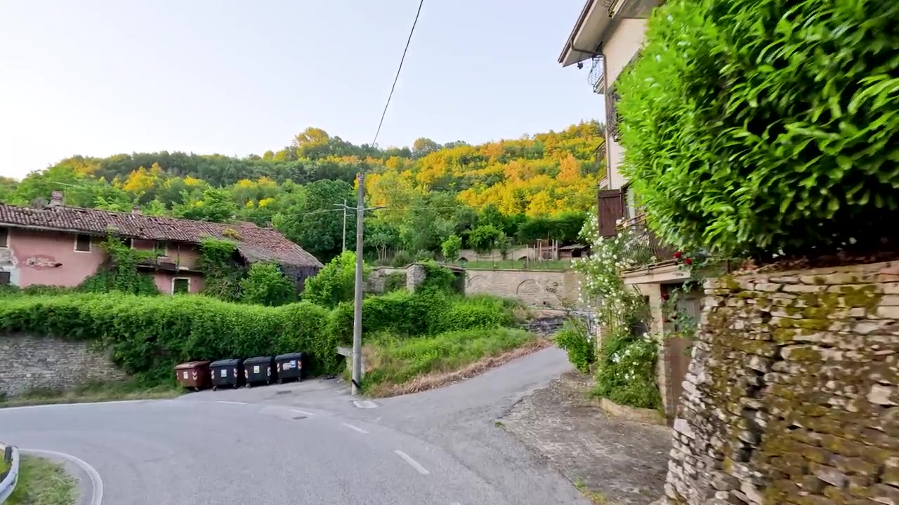 A smooth camera pan follows a curved asphalt road past stone houses and lush greenery.