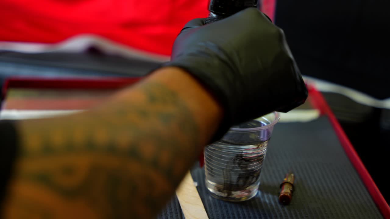 Tattoo artist gloved hand cleaning a spike of black ink in a glass of water