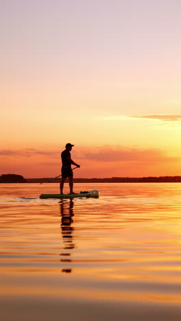 man in cap stands on the paddle board rowing with an oar. water sport practicing on the river at sunset. vertical video.
