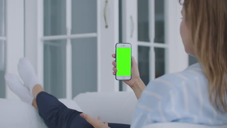 Foto-Por-Encima-Del-Hombro-De-Una-Mujer-Sosteniendo-Un-Teléfono-Móvil-O-Un-Teléfono-Inteligente-Con-Una-Pantalla-Verde-Vertical-En-La-Mano-En-Casa-Tumbada-En-El-Sofá.-La-Chica-Toca-La-Pantalla-Y-Mira-El-Contenido.