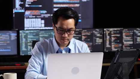 man coding at his desk in a dark room with multiple monitors