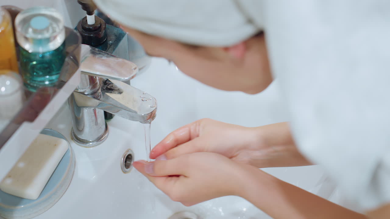 Lady gently washing her face at sink, water flowing from tap, skincare products visible beside sink, emphasizing facial cleansing and beauty routine for smooth, healthy skin