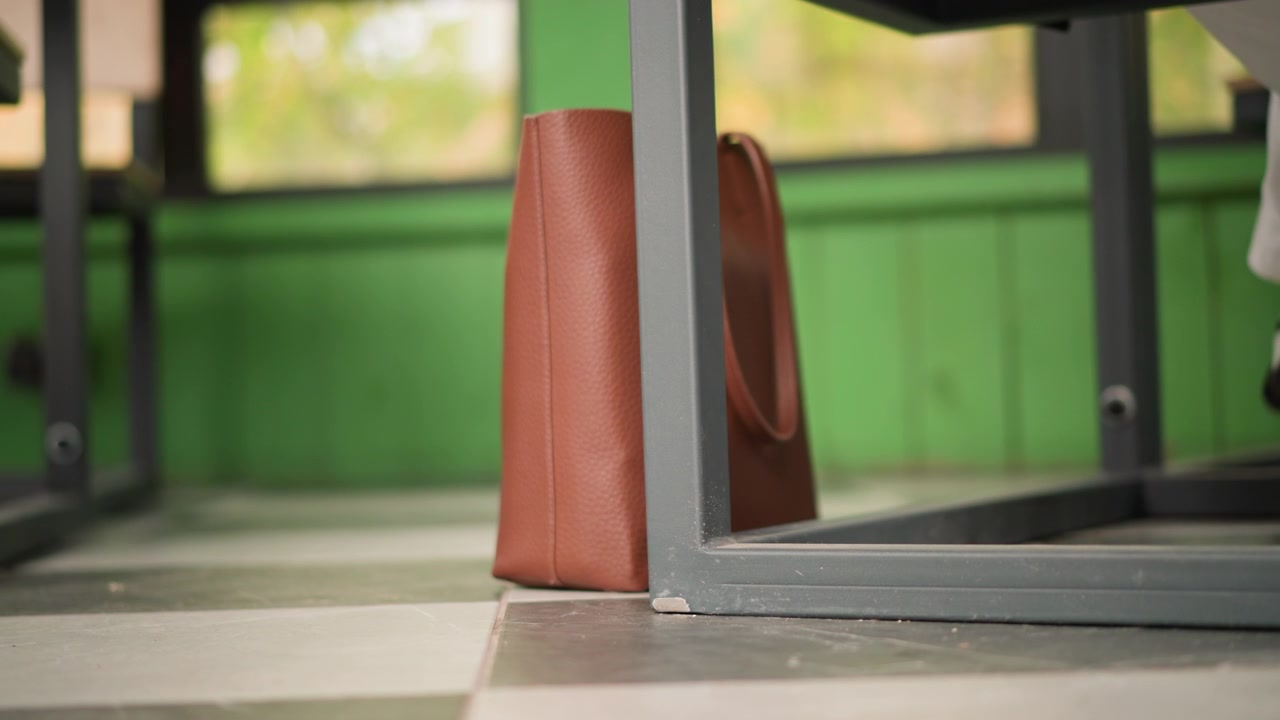 Primo piano di una borsa in pelle marrone posizionata sotto la gamba di un tavolo in un ambiente interno con pareti verdi e piastrelle a scacchiera. La scena rimane statica per tutto il tempo, concentrandosi sui dettagli della texture della borsa.