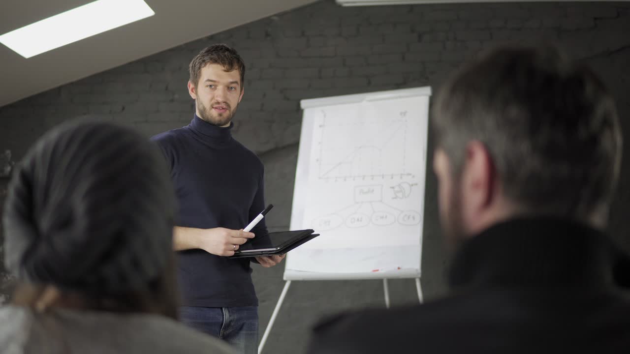 joven hombre de negocios guapo apuntando a flipchart durante la presentación en la sala de conferencias y sosteniendo la tableta.