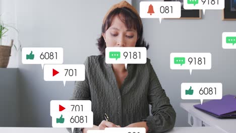 Animation-of-social-media-icons-over-asian-woman-talking-notes-and-having-a-video-call-at-office