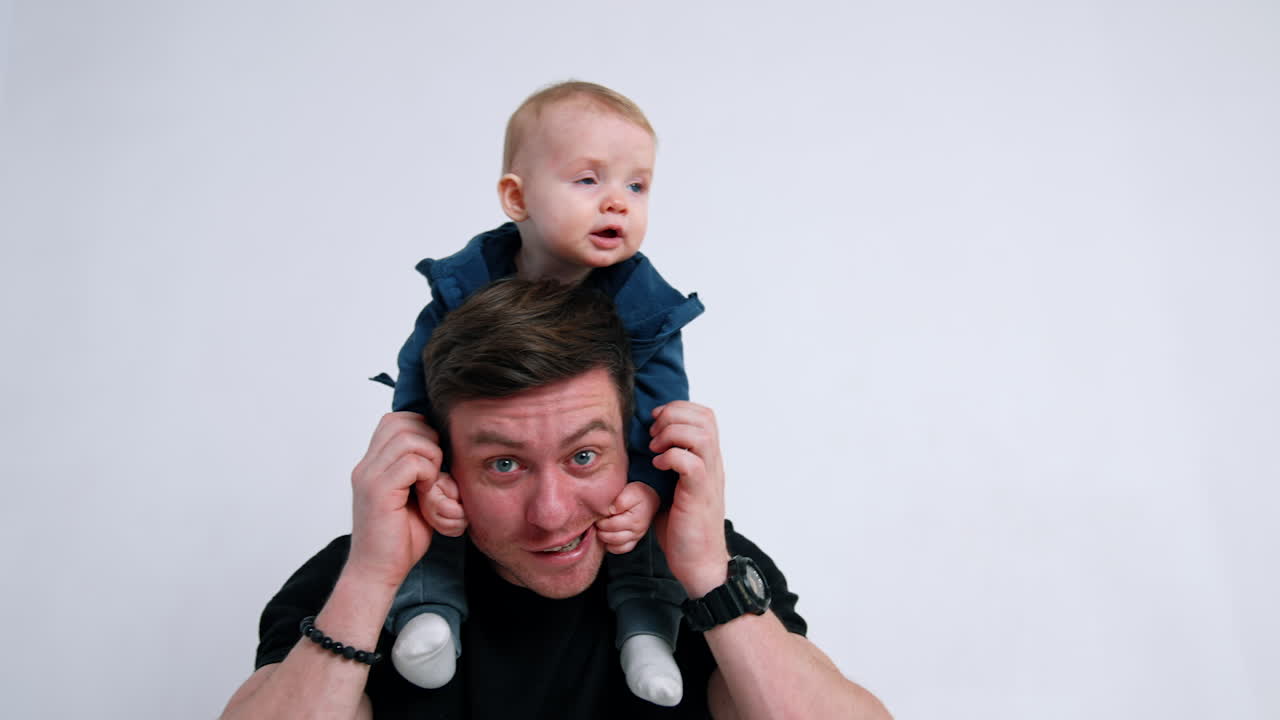 Smiling man holding a little baby on his shoulders. Cute child rubs dad's cheeks. White backdrop.